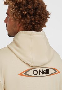 Krémová mikina s kapucňou s logom O'Neill, so vyšívanými detailmi v čiernej, oranžovej a sivej farbe, štruktúrovanou textíliou a širokou kapucňou.
