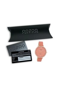 Montre OOZOO avec un cadran rond rose, des aiguilles fines et un bracelet en silicone rose clair. Comprend une carte de garantie noire et une boîte d'emballage noire.