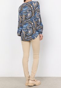 Blouse multicolore avec un motif paisley en bleu, or et blanc ; associée à un pantalon beige ajusté et des chaussures à plateforme.