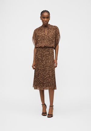 Žena stojaca v hnedých midi šatách so vzorom leopardieho kožušiny, s krátkymi rukávmi a čiernymi sandálmi na vysokom opätku s remienkom okolo členku.