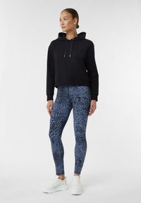 Czarna skrócona bluza z kapturem ze sznurkiem i srebrnymi końcówkami, zestawiona z niebieskimi legginsami w panterkę i białymi butami sportowymi.