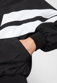Chaqueta negra con franjas diagonales blancas. Fabricada con un material ligero, con puños elásticos y una textura suave.