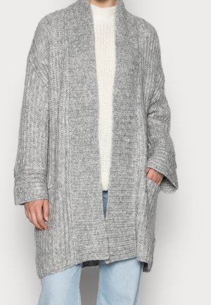 Personne portant un long cardigan en grosse maille grise sur un pull blanc et un jean bleu clair, avec une main dans la poche du cardigan.