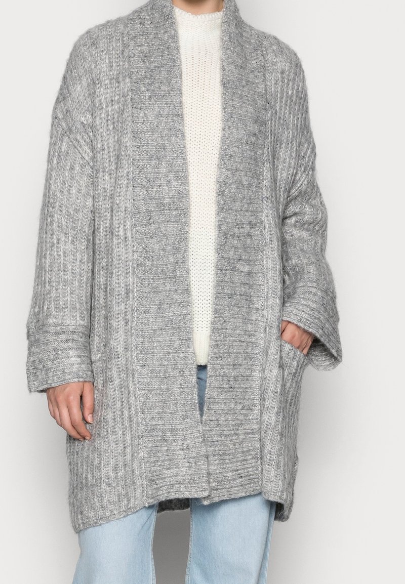 Personne portant un long cardigan en grosse maille grise sur un pull blanc et un jean bleu clair, avec une main dans la poche du cardigan.