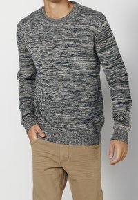 Pull en maille à motif chiné bleu marine et gris, avec un col rond et des poignets côtelés, associé à un pantalon beige.