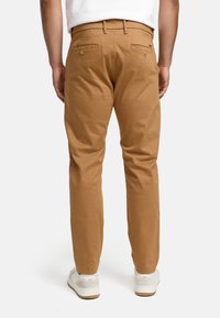 Tan cotton chinos with a slim fit, featuring two back pockets with button closures, worn with white sneakers.