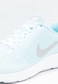 Světle modrá sportovní bota s texturovaným síťovinovým svrškem, bílými tkaničkami a stříbrným logem Nike swoosh na boku. Bílá podrážka s hladkým povrchem.