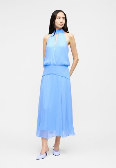 Γυναίκα με μακριά μαύρα μαλλιά, φορώντας ένα αμάνικο ανοιχτό μπλε φόρεμα με halter και ταιριαστά μυτερά τακούνια, στέκεται μπροστά σε απλό φόντο.