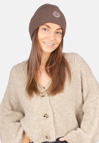 Braune gestrickte Mütze mit geripptem Muster und einem Smiley-Emblem, kombiniert mit einem hellbeigen, grob gestrickten Pullover mit dekorativen Knöpfen.