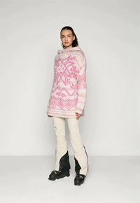 Γυναίκα που φοράει μια ροζ και λευκή πουλόβερ με σχέδιο, μπεζ παντελόνι με μαύρους κάτω πίνακες και μαύρες χειμερινές μπότες, στέκεται μπροστά σε έναν απλό τοίχο.