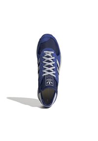 Modrá sportovní bota vyrobená z textilu a semiše, s bílou podrážkou, šedými tkaničkami a třemi podpisovými pruhy na boku.