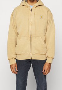Sudadera con cremallera color beige hecha de una tela suave, que cuenta con un bolsillo frontal, puños acanalados y un parche con el logo en el lado izquierdo del pecho.