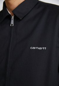 Czarna kurtka z zamkiem błyskawicznym, wykonana z trwałego materiału, z powiększonym kołnierzem. Posiada srebrny zamek i haftowane białe logo "carhartt" na piersi.
