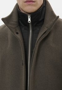 Mand iført en mørkebrun uldfrakke over en sort zip-up jakke med høj krave, vist fra læberne til midt på brystet.