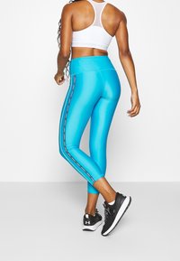 Niebieskie legginsy w wysokim stanie z błyszczącym wykończeniem i czarnymi bocznymi paskami, w połączeniu z czarnymi butami sportowymi, prezentujące dopasowany krój.