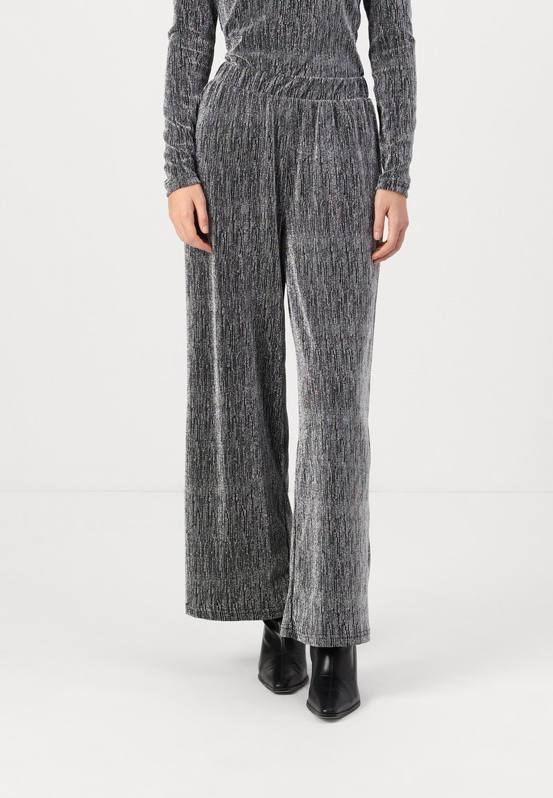 Pantalons à larges jambes en tissu gris texturé avec des fils métalliques. Taille haute avec des plis subtils, associés à des bottines noires.