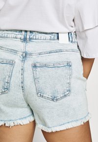 Vaaleansiniset farkkushortsit, joissa on rispaantunut reuna, kaksi takataskua ja valkoinen merkki vyötäröllä. Kankaan pinta on teksturoitu ja haalistunut.