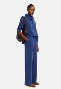 Niebieska, luźna bluza z wysokim kołnierzem i zapięciem na guziki, w połączeniu z szerokimi spodniami w pasującym kolorze oraz brązowymi butami wsuwanymi.