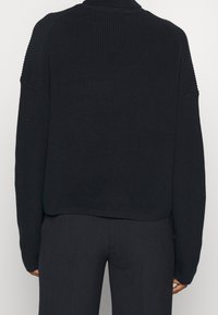 Persona che indossa un maglione a coste nero e pantaloni scuri, vista da dietro, in piedi contro uno sfondo chiaro e uniforme.