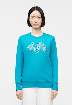 Junge Frau mit glatten schwarzen Haaren trägt ein leuchtend blaues Versace Jeans Couture Sweatshirt und weiße Hosen, steht vor einem schlichten weißen Hintergrund.