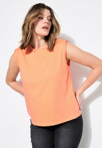 Mouwloos oranje top van zachte katoen met een ronde halslijn, losse pasvorm en zijsplitten, gecombineerd met donkere jeans voor contrast.