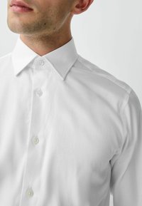 Weißes Hemd mit Knopfleiste, klassischem Kragen, glatter Textur und zwei sichtbaren Knöpfen an der Knopfleiste. Sauberes, minimalistisches Design.