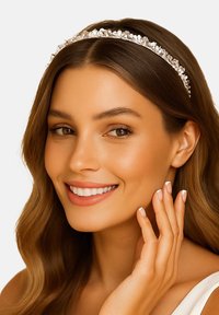 Opaska na głowę zdobiona kryształami na srebrnym tle; gładkie włosy opadają na ramiona, ręka delikatnie dotyka twarzy, neutralne tło.