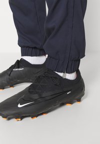 Schwarze Fußballschuhe mit strukturiertem Obermaterial, orangefarbenen Noppen und weißem Nike-Swoosh. Kombiniert mit taillierten, marineblauen Hosen und weißen Socken.