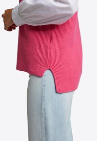 Rosa gestricktes ärmelloses Oberteil mit gerippter Textur, seitlichen Schlitzen und einem asymmetrischen Saum, getragen über hellblauen Jeans.