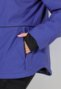 Lila vattentät jacka med svarta accenter, utrustad med en sidoficka och elastiska ärmslut. Släta, lätta tyger med en modern passform.