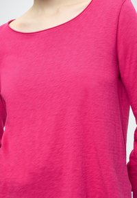Dolga roza majica z dolgimi rokavi iz lahke, teksturirane tkanine s širokim izrezom. Ponuja subtilne odtenke barve in sproščen kroj.