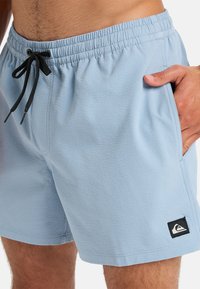 Light blue swim shorts with a textured fabric, elastic waistband, black drawstring, and side pockets. Features a small black logo patch.