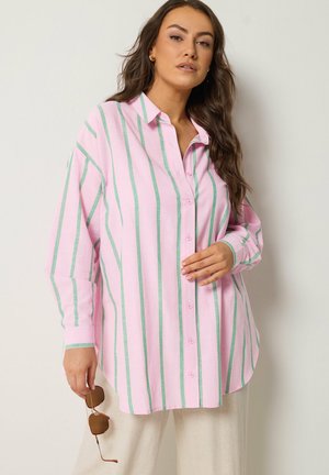 Kvinde iført løs lyserød skjorte med lodrette grønne striber, holder brune solbriller i venstre hånd, stående mod en ensfarvet lys væg.