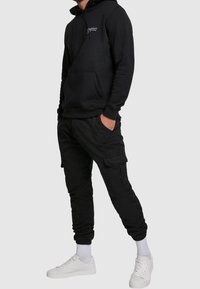 Felpa nera con cappuccio, tasca frontale e logo, abbinata a pantaloni cargo neri con tasche. Indossata con sneakers bianche e calzini.