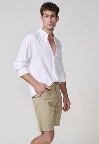 Cămașă albă ușoară cu un croi lejer, asortată cu pantaloni scurți bej ce au buzunare laterale, față plată și o textură netedă.