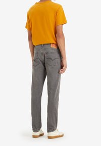 Camiseta amarilla de manga corta combinada con jeans rectos grises, con un parche de cuero en la parte posterior y detalles de costura sutiles. Zapatillas blancas.