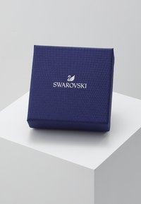 Tumši zils, teksturēts dāvanu kastīte ar magnētisku aizdari, uz kuras priekšpuses ir sudraba Swarovski logotips. Kvadrāta forma ar gludiem maliem.