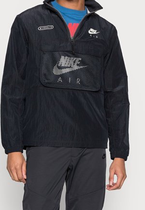 Person iført en sort Nike Air vindjakke med netlommer foran og logoer, en blå og rød skjorte under, og sorte bukser.