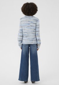 Kortsukurkse juustega inimene kannab sinist ja halli triibulist sviitrit ning laiadest sinistest teksades, seistes seljaga kaamera poole tasase tausta ees.