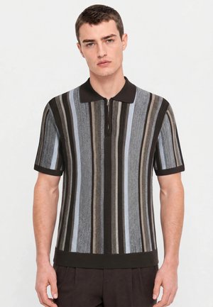 Jeune homme portant un polo en maille à manches courtes avec des rayures verticales en noir, gris, bleu et marron sur un fond uni.