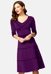 Valkoinen mekko, jossa on V-kaula-aukko ja kolmiosaiset hihat. Mekossa on kerrostettu muotoilu ja vaakasaumat ympäri hameen. Sileä rakenne.