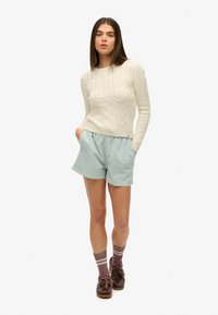 Crème kabelgebreide trui, gestreepte mintgroene shorts, bruine schoenen en grijze sokken met witte strepen. De nadruk ligt op texturen en zachte kleuren.