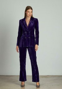 Blazer doppiopetto in velluto viola abbinato a pantaloni coordinati. Presenta linee verticali, revers e bottoni dorati per un tocco di eleganza.
