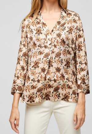 Femme portant un chemisier beige à fleurs à manches longues avec col en V et un pantalon couleur crème, debout devant un fond uni.