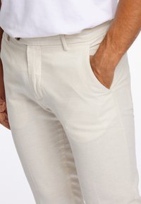 Mężczyzna w jasnobeżowych spodniach z ręką w prawej kieszeni, w połączeniu z białą koszulą na jednolitym tle.