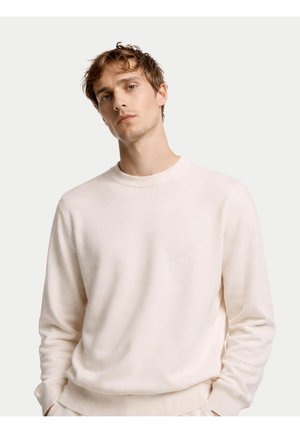 Junger Mann mit zerzaustem Haar, der einen schlichten weißen Langarmpullover trägt und mit den Händen in den Taschen vor hellem Hintergrund steht.