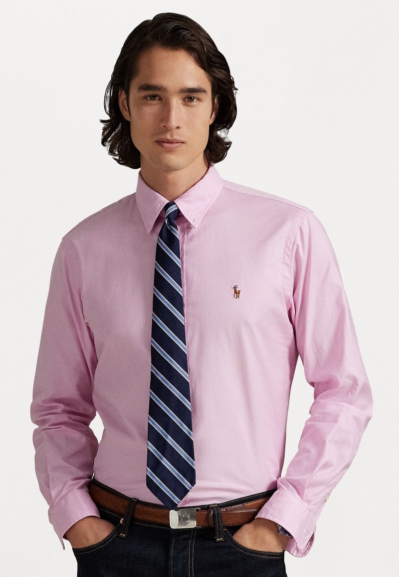 Camicia button-up rosa chiaro con maniche lunghe, caratterizzata da un piccolo logo sul petto e una cravatta a righe blu navy. Jeans scuri e cintura marrone visibili.