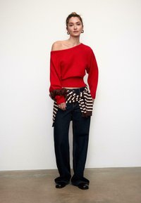 Roter Schulterfreier Pullover mit gerippten Bündchen, kombiniert mit hoch taillierter dunkler Jeans. Braun-weiß gestreifter Stoff um die Taille gebunden, schwarze Schuhe.