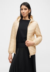 Γυναίκα που φοράει μπεζ μπουφάν με καπιτονέ σχέδιο πάνω από μαύρο τοπ και μαύρη πλισέ φούστα, κρατώντας το φερμουάρ του μπουφάν με το ένα χέρι.