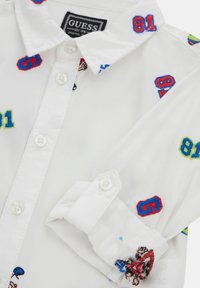 Witte shirt met kleurrijke geborduurde cijfers en een ontwerp, met een knoopsluiting bij de kraag en manchetten gemaakt van een gladde stof.